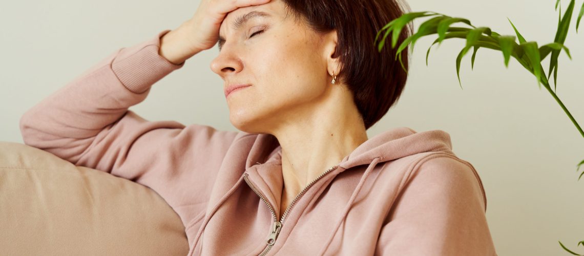Woman putting hand on head due to headache after waking up, filling deep sadness. Migraine. Mental health of mature female, generation X. Tired lady sitting in home with closed eyes